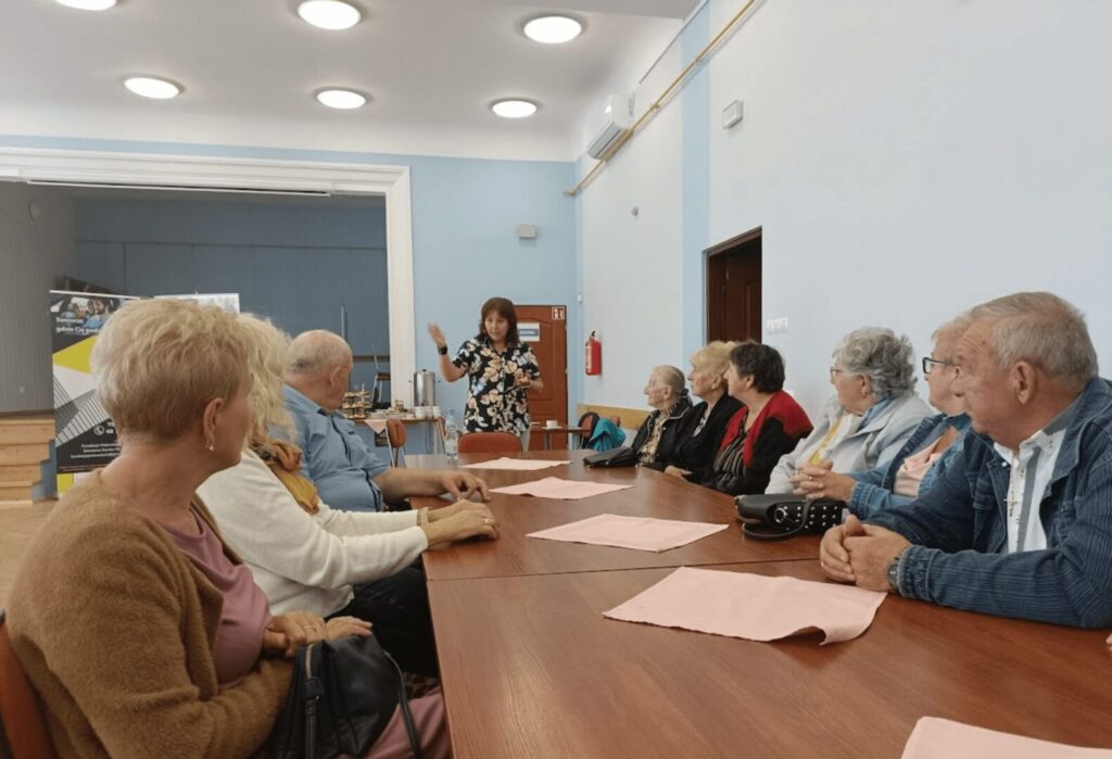 Grupa seniorów siedzi przy stole, słuchając kobiety prowadzącej spotkanie w jasnej sali, na stole leżą różowe serwetki.
