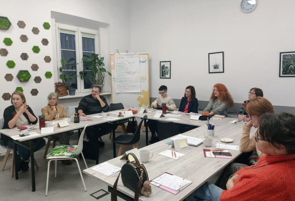 Grupa osób siedzi przy stołach w jasnej sali podczas spotkania, rozmawiają i notują; na ścianach zielone dekoracje i rośliny w ramkach.