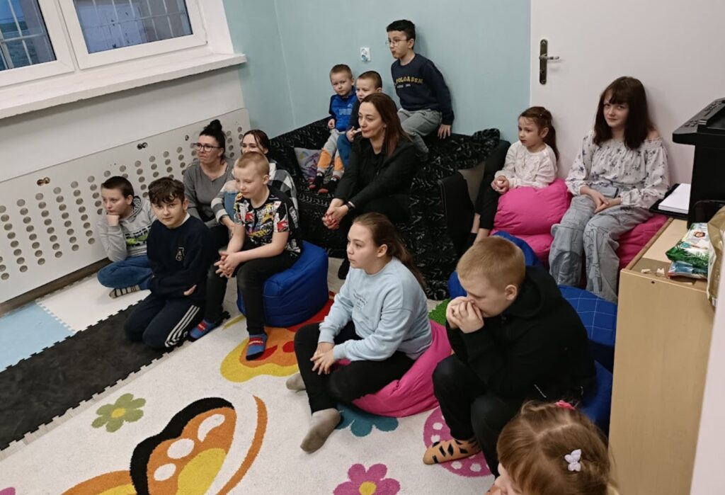 Grupa dzieci i opiekunów siedzi na pufach i dywanie w kolorowej sali, wspólnie oglądają coś poza kadrem, panuje skupiona atmosfera.