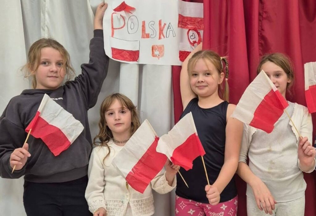 Cztery dziewczynki trzymają biało-czerwone flagi i stoją na tle dekoracji patriotycznej z napisem "Polska" i rysunkami flag.