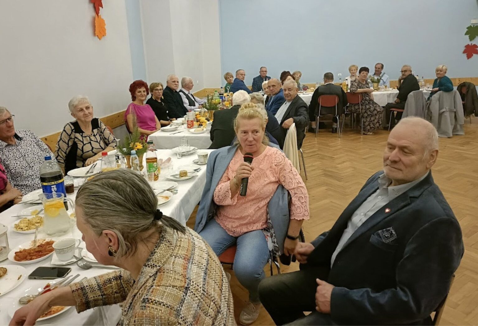 Grupa seniorów siedzi przy stołach podczas spotkania towarzyskiego; jedna z kobiet trzyma mikrofon i zabiera głos, w tle dekoracje jesienne.