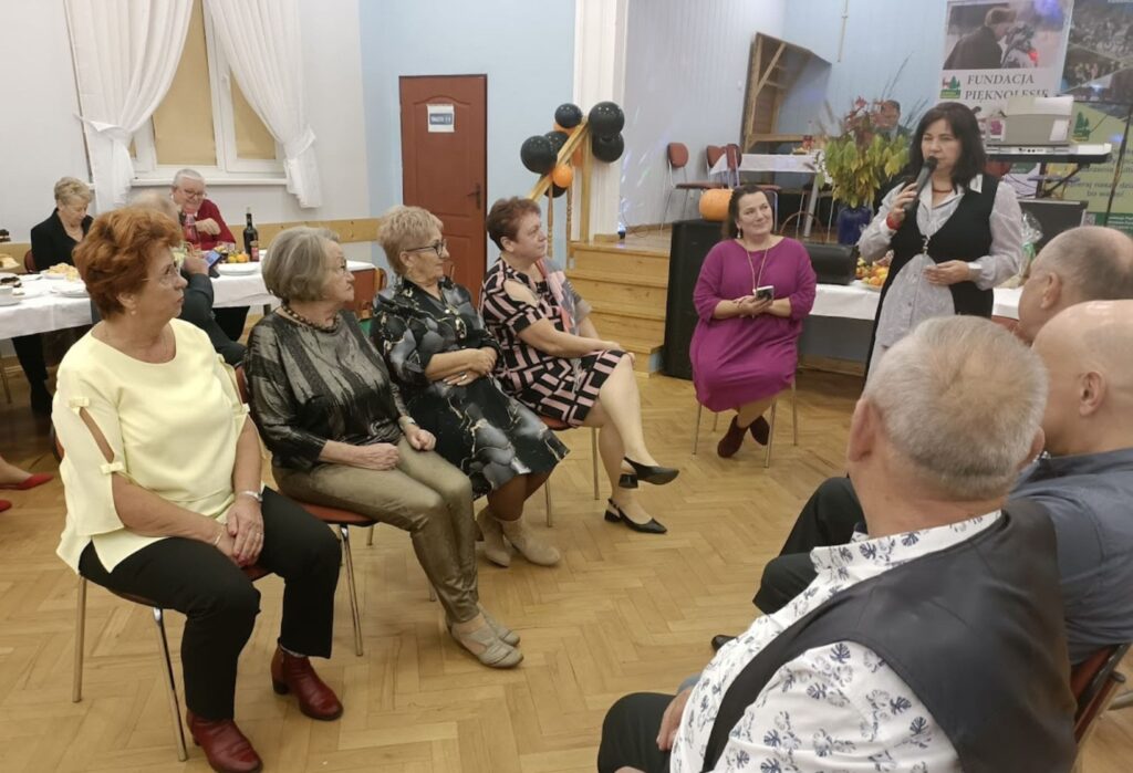 Grupa seniorów siedzi w półokręgu na sali, jedna kobieta stoi z mikrofonem i przemawia, w tle widać balony, stół z poczęstunkiem i baner Fundacji Pięknolesie.