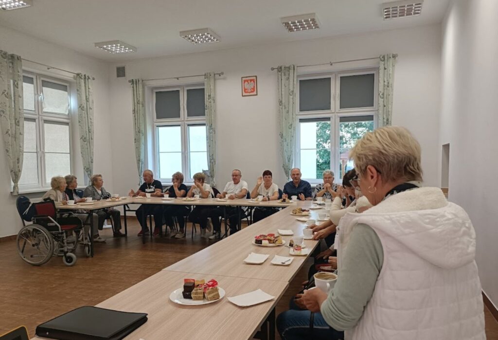 Seniorzy siedzą przy stołach ustawionych w podkowę, piją kawę i jedzą ciasto podczas spotkania w jasnej sali z dużymi oknami.