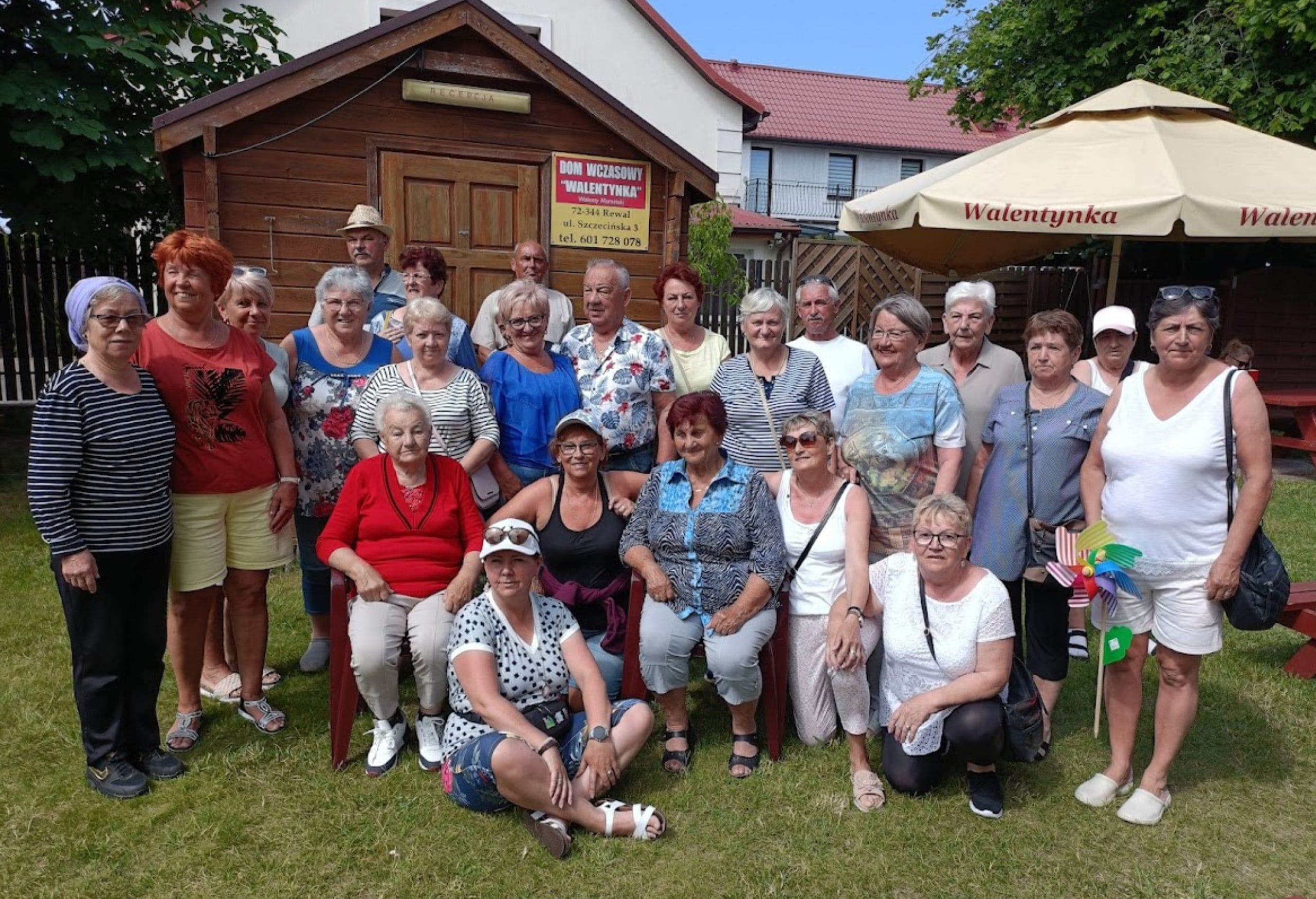 Grupa seniorek i seniorów pozuje do wspólnego zdjęcia na trawie przed drewnianym domkiem z szyldem "Dom Wczasowy Walentynka", obok widoczne parasole ogrodowe.