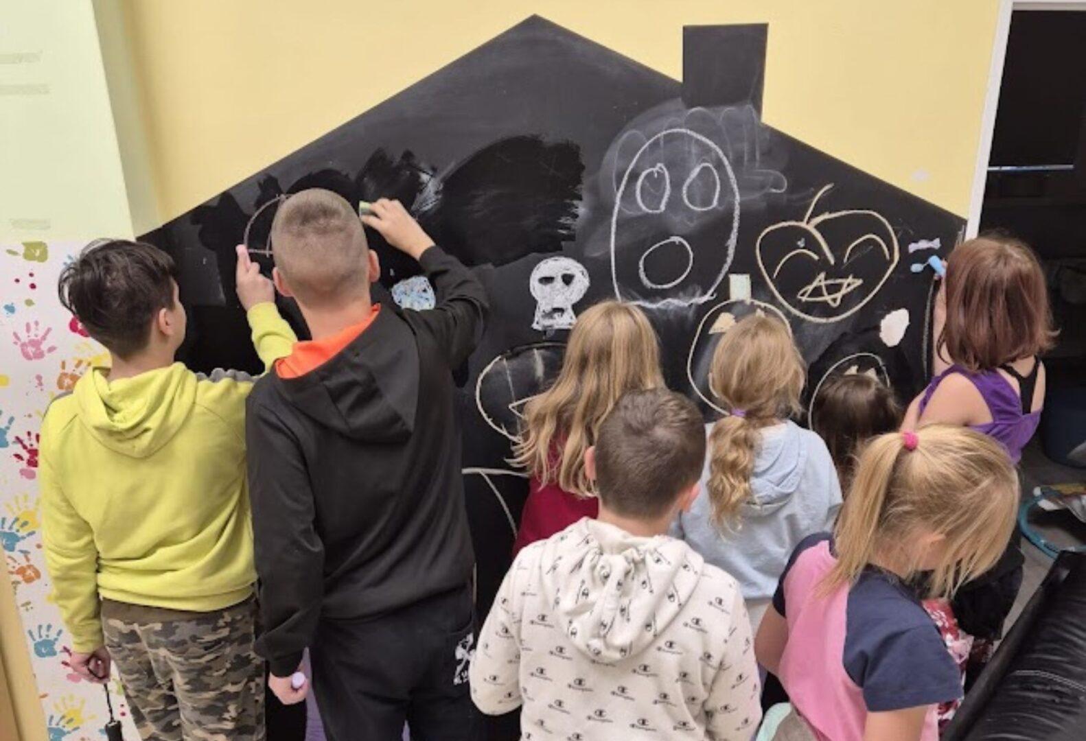 Grupa dzieci rysuje kredą na dużej czarnej tablicy w kształcie domu, tworząc różne obrazki.