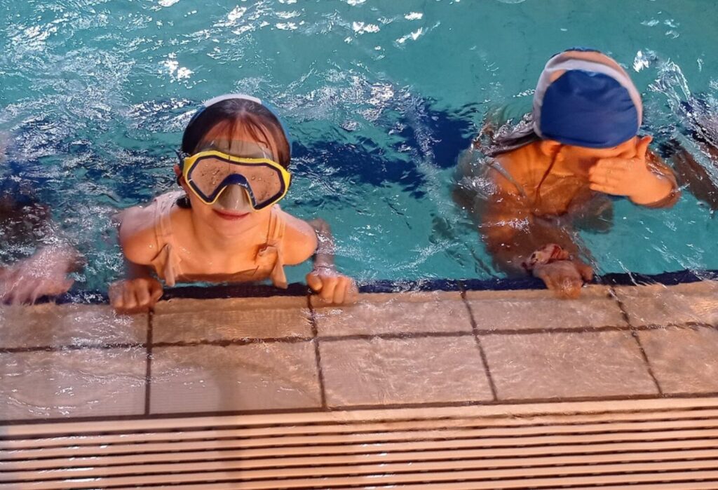 Dwoje dzieci w basenie trzyma się krawędzi, jedno ma na sobie maskę do nurkowania, drugie zakrywa twarz.