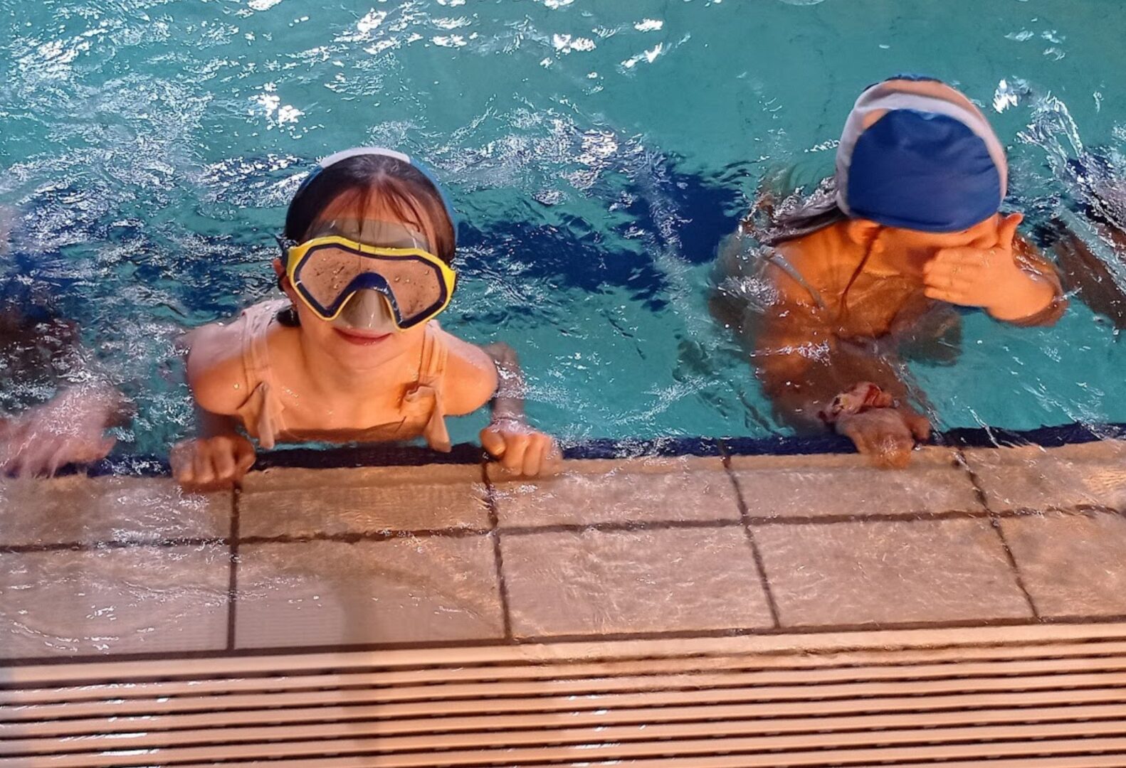 Dwoje dzieci w basenie trzyma się krawędzi, jedno ma na sobie maskę do nurkowania, drugie zakrywa twarz.