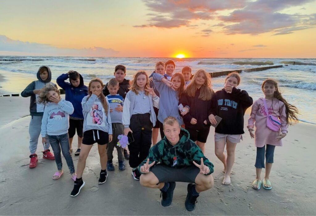 Grupa dzieci pozuje na plaży o zachodzie słońca, w tle morze i kolorowe niebo.