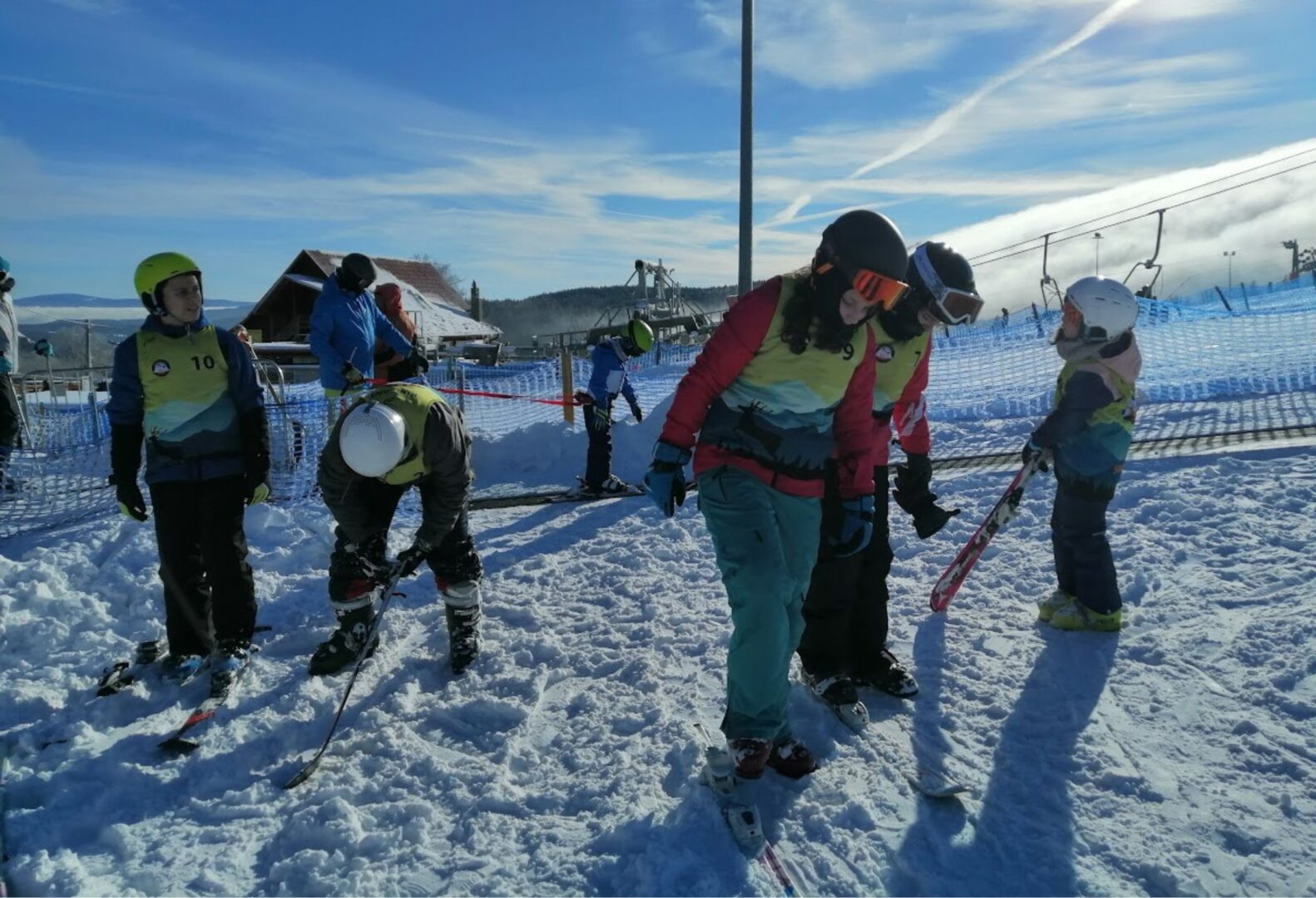 Dzieci i instruktorzy na stoku narciarskim, przygotowują się do jazdy na nartach; śnieżne otoczenie, niebieskie niebo i wyciąg narciarski w tle.