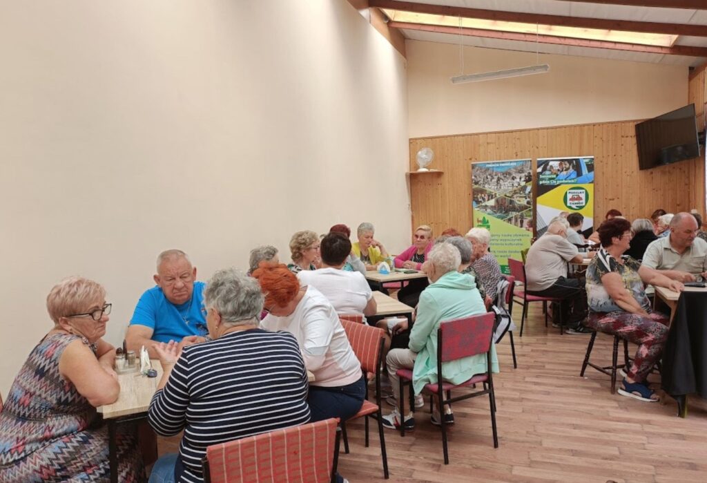 Grupa seniorów siedzi przy stołach w sali i rozmawia w parach, w tle widoczne są plakaty informacyjne i drewniana ściana.