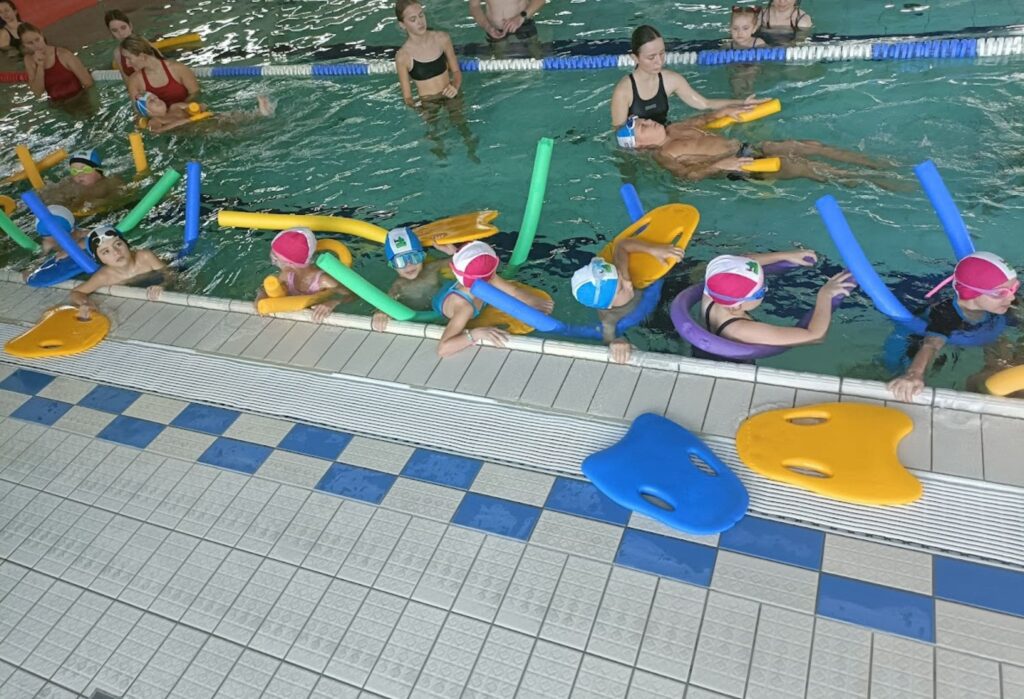 Dzieci w czepkach i z kolorowymi makaronami uczą się pływać w basenie, przy krawędzi stoi instruktorka, a w tle widać opiekunów.