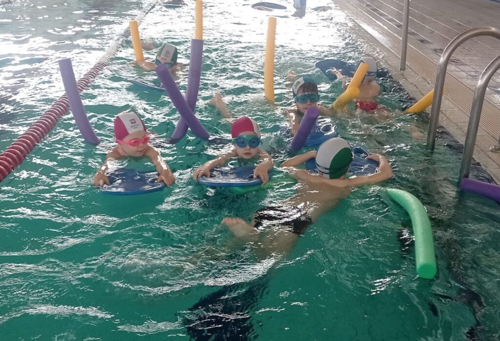 Dzieci uczące się pływać z pomocą desek i makaronów w basenie krytym.