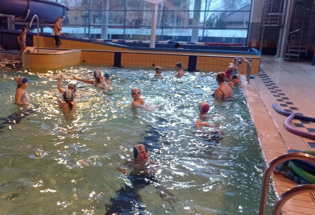 Dzieci w czepkach pływają w basenie pod okiem instruktorów.