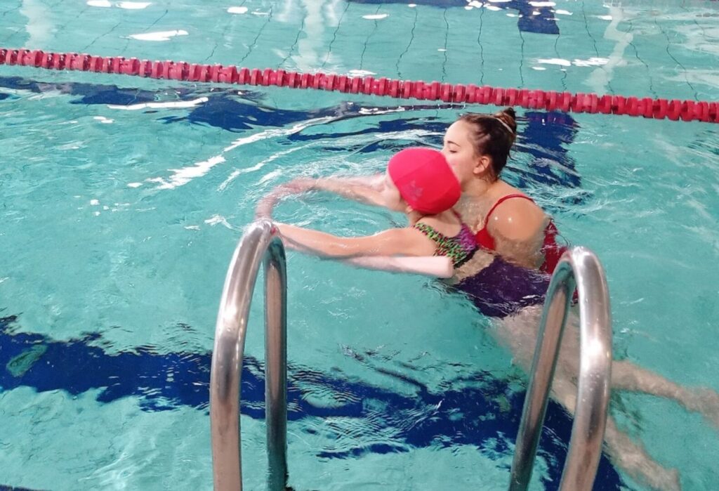 Instruktorka uczy dziecko w czerwonym czepku pływać na basenie.