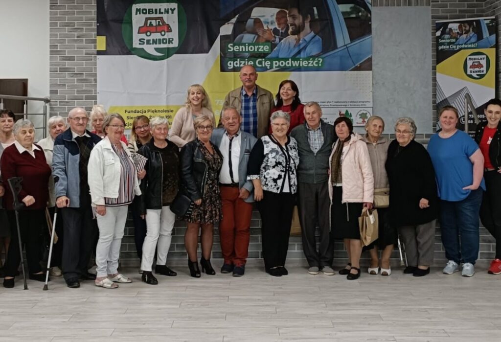Grupa seniorów i organizatorów pozuje do wspólnego zdjęcia na tle plakatu promującego projekt „Mobilny Senior” z hasłem „Seniorze, gdzie Cię podwieźć?”.