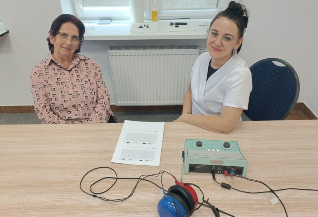Starsza kobieta i młoda specjalistka siedzą przy stole; na stole leży aparat do badania słuchu, słuchawki i karta wyników.