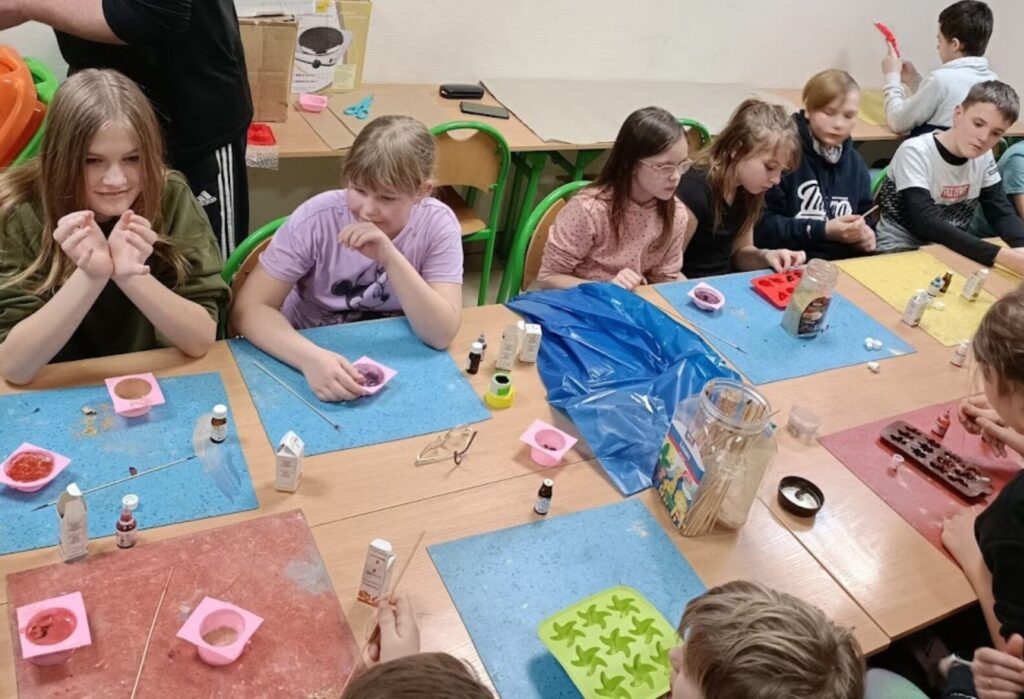 Grupa dzieci siedzi przy stole i tworzy rękodzieło podczas warsztatów; na stole kolorowe podkładki, foremki i materiały do pracy.
