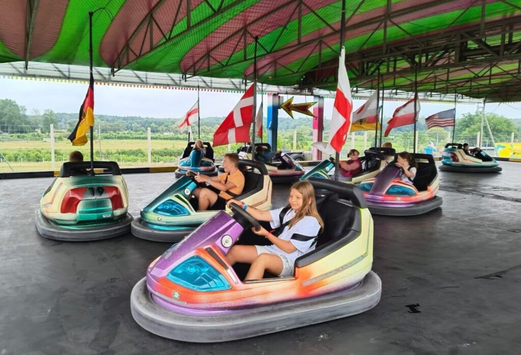 Dzieci jeżdżą w kolorowych samochodzikach elektrycznych na autodromie, nad nimi powiewają flagi różnych państw.