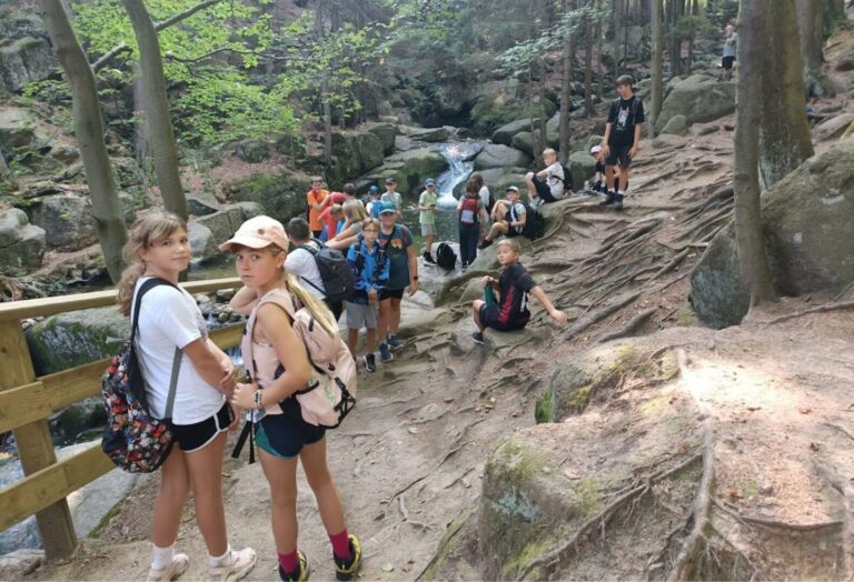 Grupa dzieci i młodzieży podczas wycieczki górskiej, wędrująca leśnym szlakiem w pobliżu strumienia i skał.