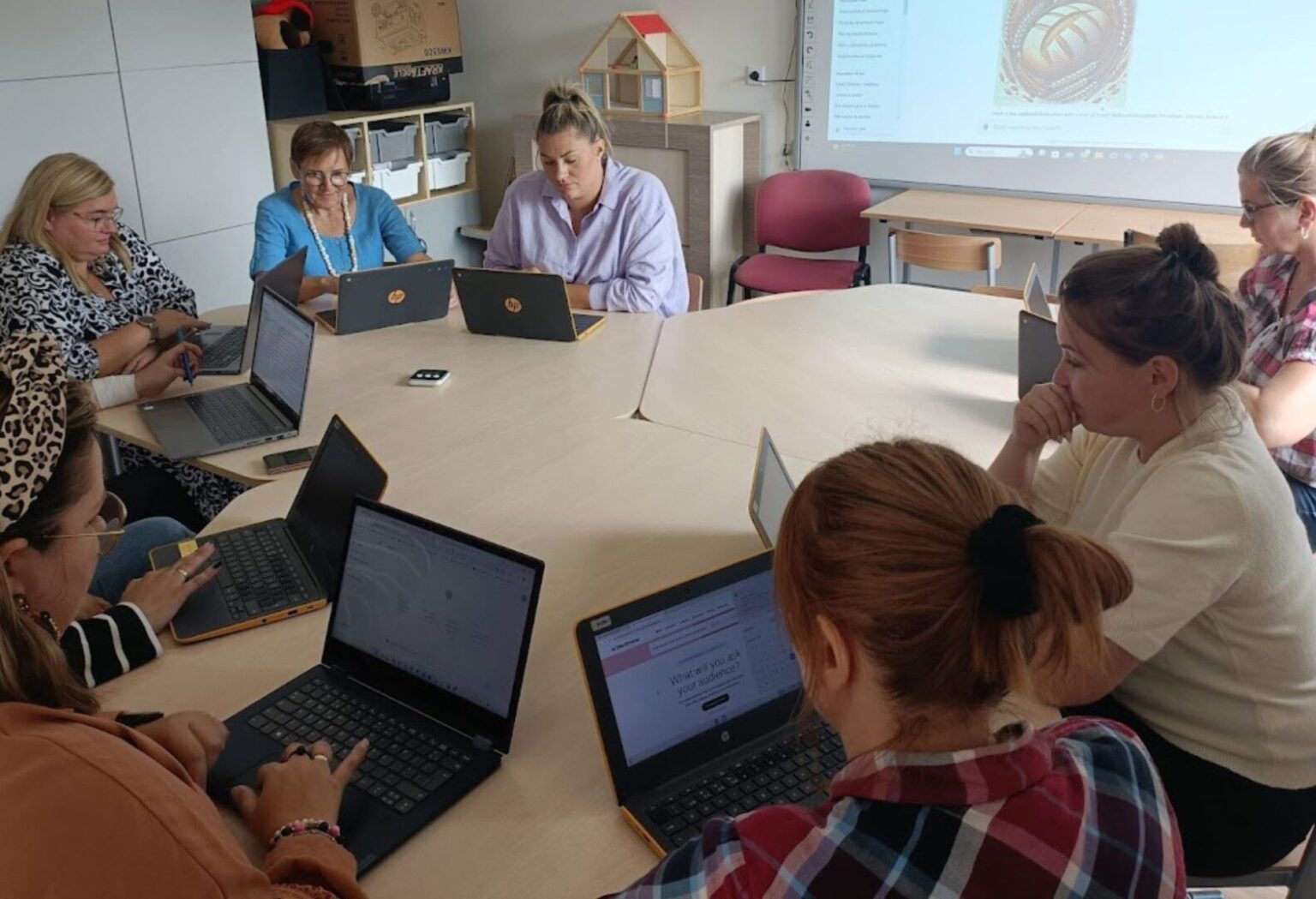 Grupa kobiet siedzi przy okrągłym stole, każda z laptopem, uczestnicząc w szkoleniu komputerowym; w tle widoczna prezentacja na rzutniku.
