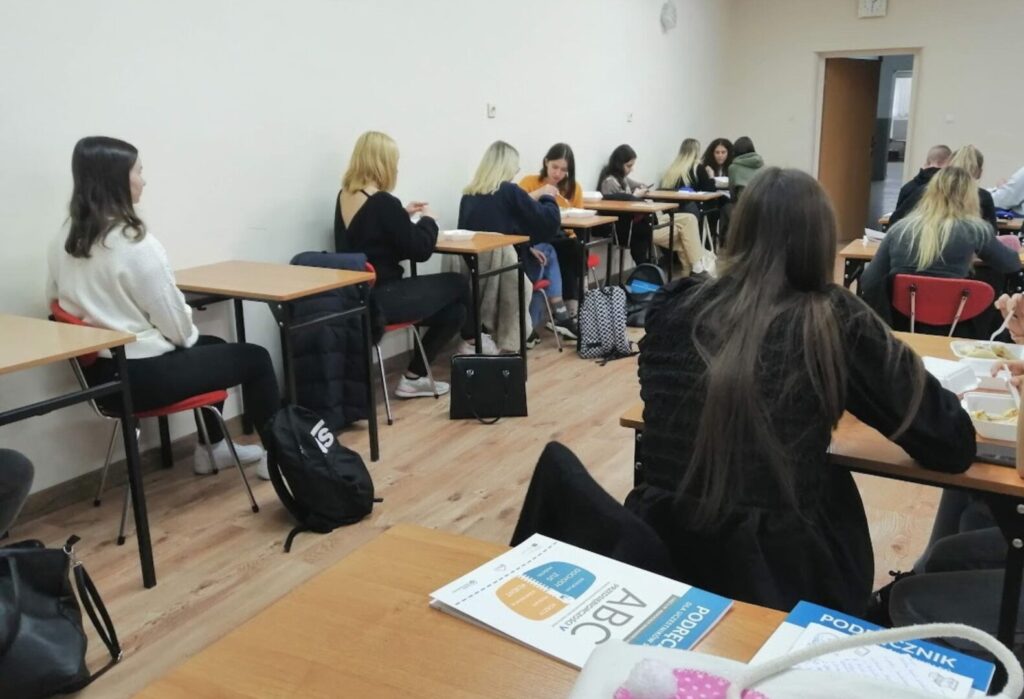 Młodzież siedząca przy ławkach w sali lekcyjnej, zajęta pracą z materiałami edukacyjnymi.