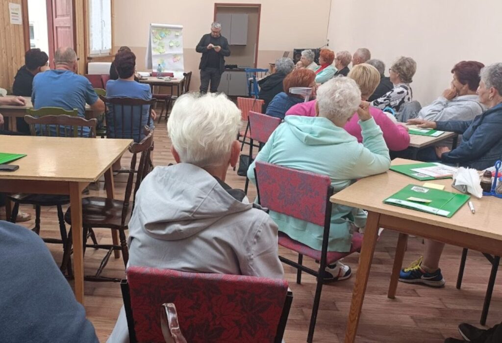 Seniorzy uczestniczący w szkoleniu i spotkaniu integracyjnym, siedzący przy stołach w sali.