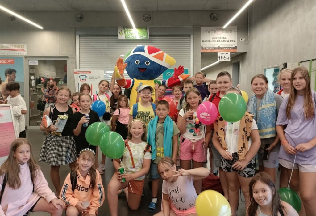 Grupa uśmiechniętych dzieci pozuje do zdjęcia z kolorową maskotką w holu budynku; część dzieci trzyma balony i napoje, niektóre mają ręczniki na szyjach – prawdopodobnie po zajęciach na basenie.