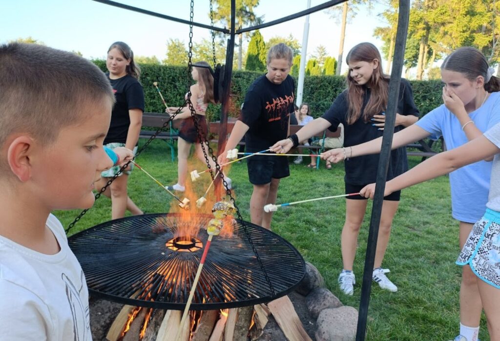 Grupa dzieci piecze kiełbaski i pianki nad ogniskiem w ogrodzie; panuje radosna atmosfera letniego wieczoru.