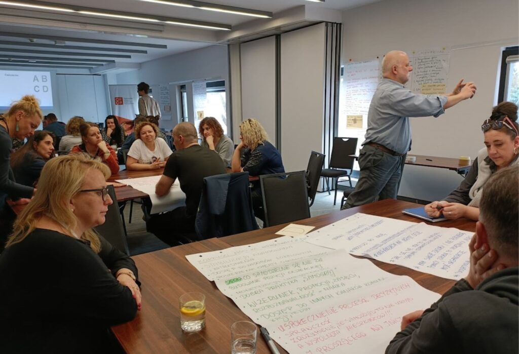 Grupa osób uczestniczy w warsztatach – siedzą przy stołach z arkuszami papieru i notują, a prowadzący mężczyzna stoi przy tablicy i gestykuluje.