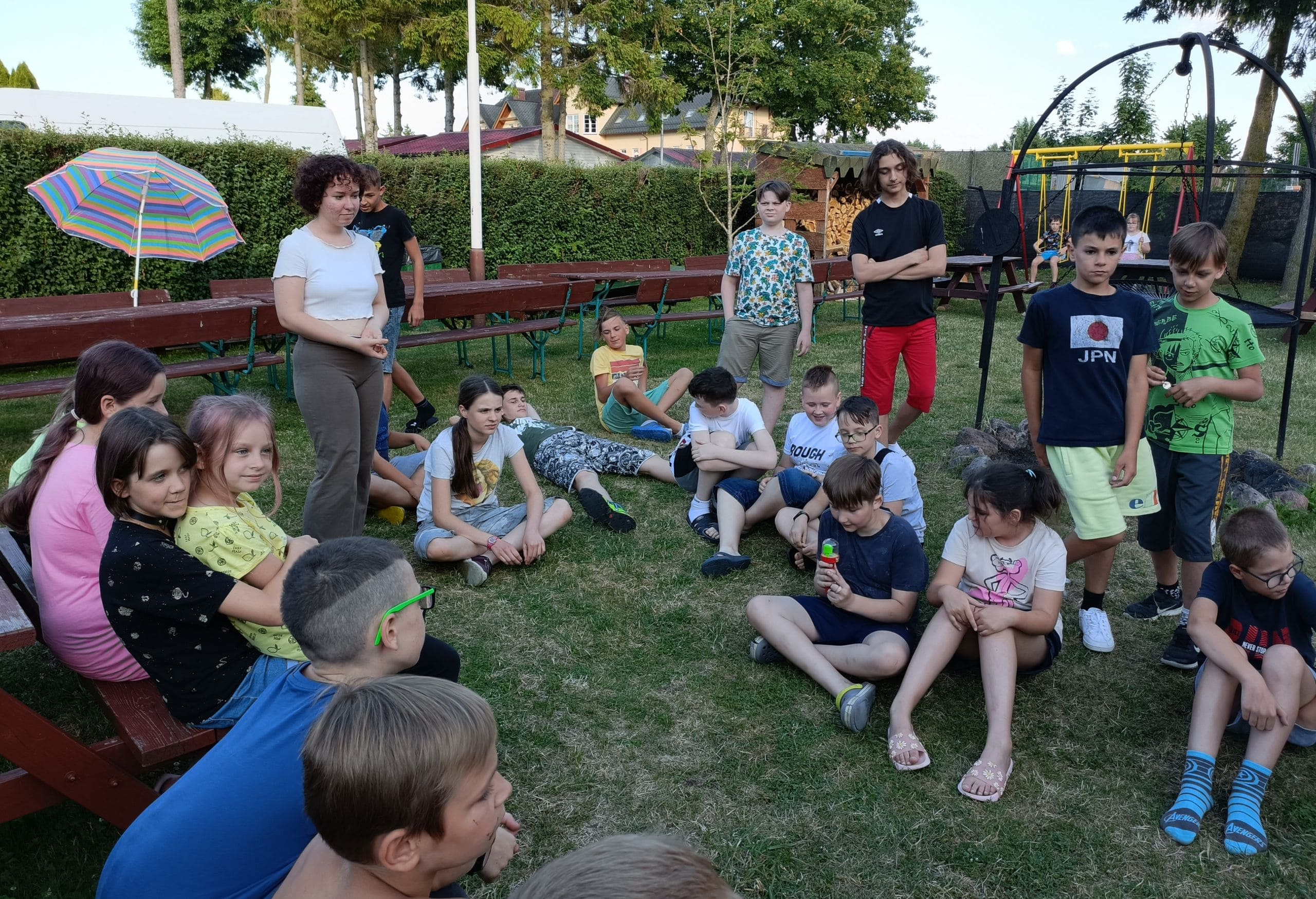 Dzieci siedzące i stojące w kręgu na trawie, podczas wspólnej rozmowy lub gry integracyjnej na świeżym powietrzu; obok animatorka.