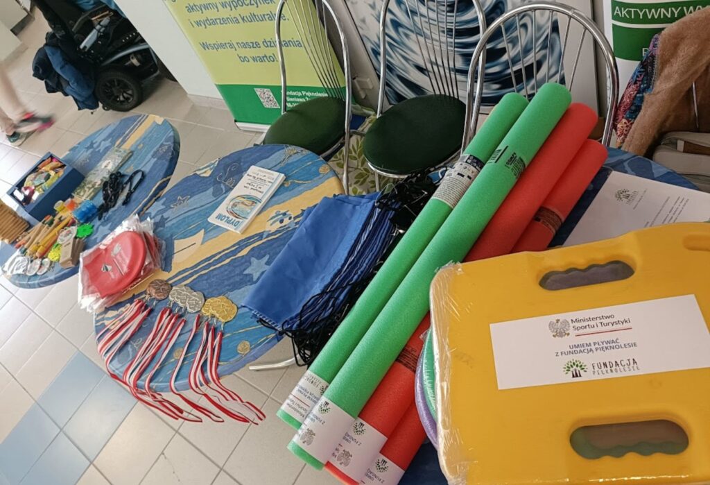 Stół z materiałami do nauki pływania – piankowe makarony, deski, czepki, medale i torby sportowe, obok ulotki i informacja o dofinansowaniu z Ministerstwa Sportu i Fundacji Pięknolesie.