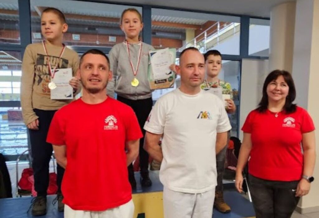 Dzieci z medalami stoją na podium z dyplomami w rękach, przed nimi dwoje trenerów i kobieta w czerwonej koszulce – wszyscy uśmiechnięci.