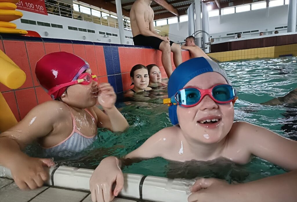 Dwoje dzieci w czepkach i okularach pływackich trzyma się brzegu basenu, uśmiechają się i rozmawiają; w tle inne osoby pływają lub siedzą przy brzegu.