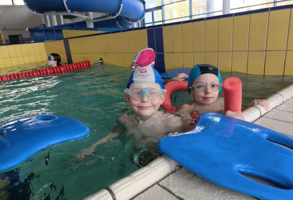 Dwoje dzieci w czepkach i okularach pływackich trzyma się brzegu basenu z deskami i piankowymi makaronami, w tle zjeżdżalnia wodna.