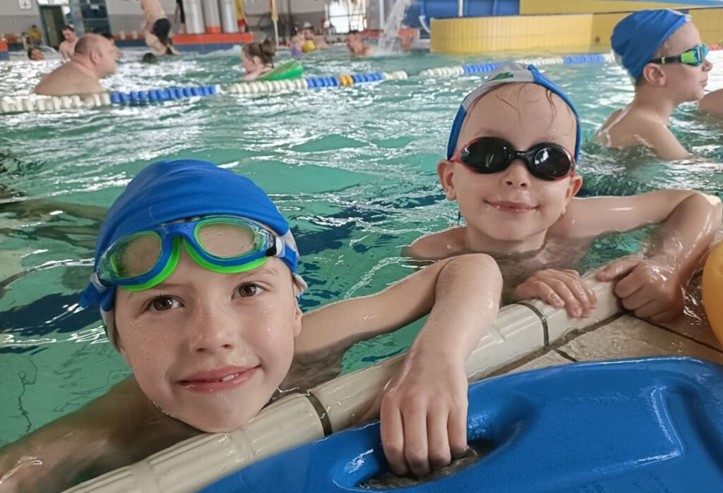 Dwóch uśmiechniętych chłopców w czepkach i okularach pływackich trzyma się brzegu basenu podczas nauki pływania.