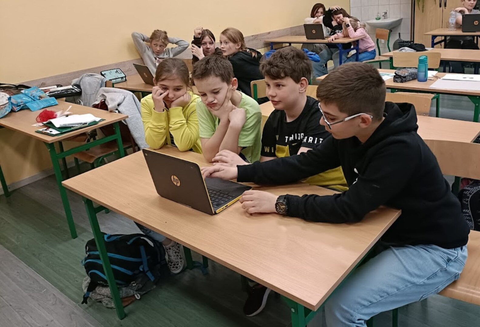 Grupa dzieci siedzi przy szkolnej ławce, wspólnie pracując na laptopie, w tle inne grupy również zaangażowane w zajęcia.