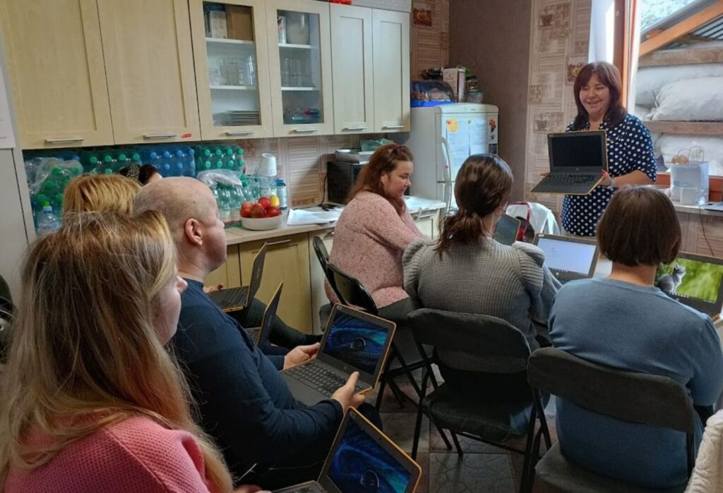 Grupa dorosłych siedzi w kuchni podczas szkolenia z laptopami, kobieta prowadząca prezentuje coś na ekranie komputera.