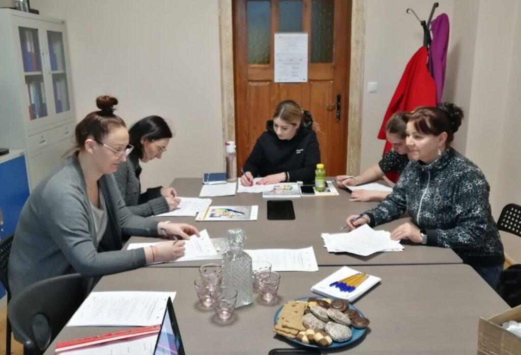 Pięć kobiet siedzi przy stole, uczestnicząc w szkoleniu z materiałami papierowymi, na stole znajdują się również napoje i przekąski.