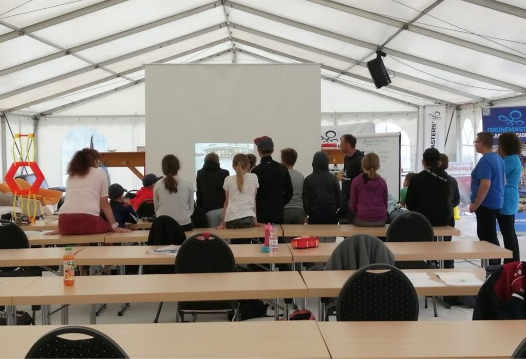 Grupa osób ogląda prezentację na dużym ekranie w namiocie szkoleniowym; na pierwszym planie rzędy pustych krzeseł i stołów.