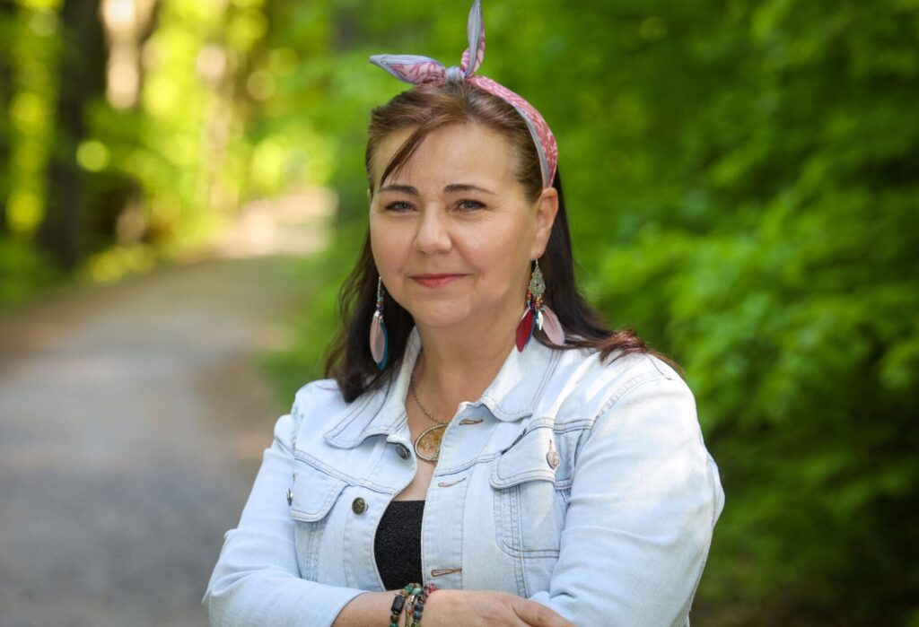 Krystyna Adamenko uśmiecha się stojąc na leśnej ścieżce, ubrana w jasną kurtkę jeansową i opaskę we włosach.