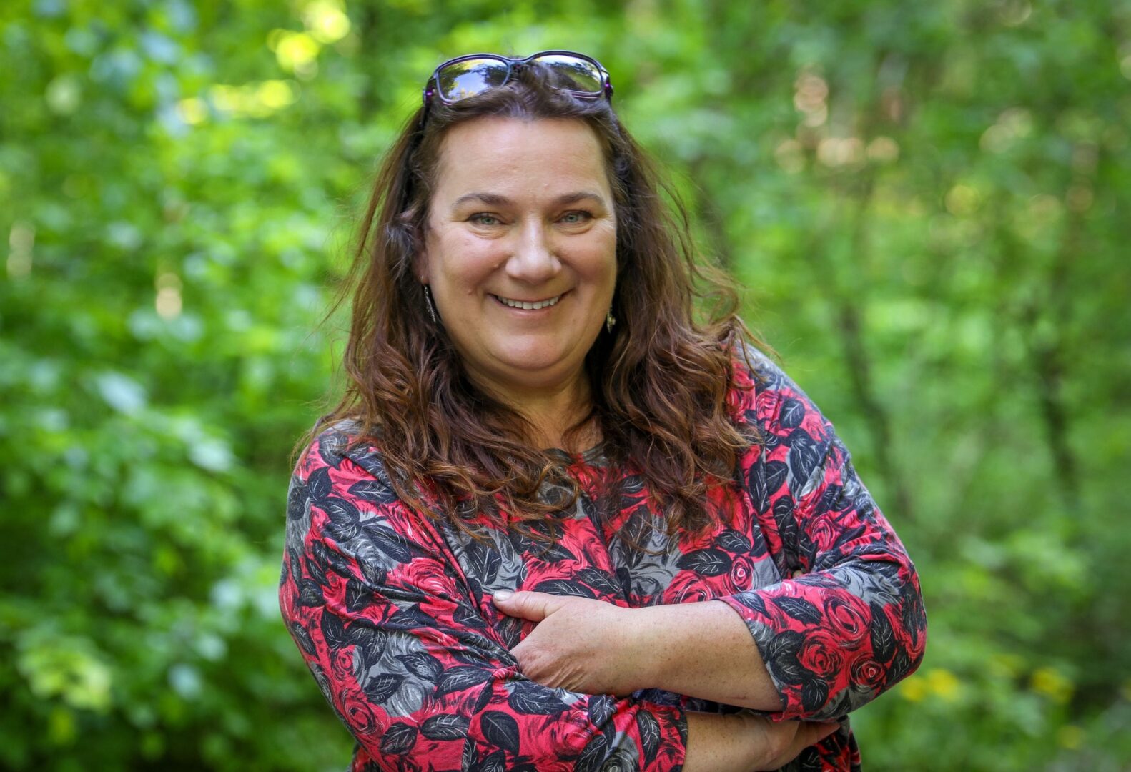 Monika Sznajder uśmiecha się pogodnie, ubrana w kolorową sukienkę, stoi na tle zieleni lasu.