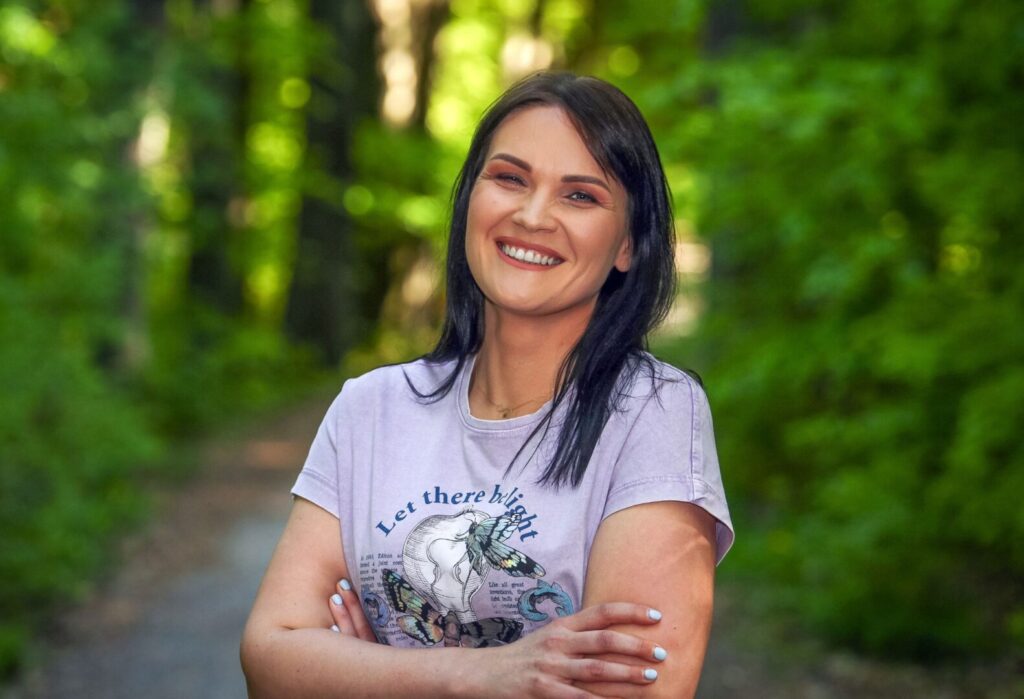 Katarzyna Ufa uśmiecha się serdecznie, stojąc na tle zielonego lasu, ubrana w liliowy T-shirt z nadrukiem.
