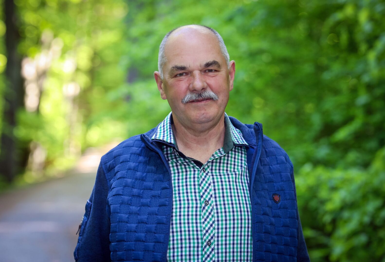 Bogusław Gołojuch uśmiecha się łagodnie, ubrany w kraciastą koszulę i granatową kamizelkę, stoi na tle zieleni leśnej alejki.