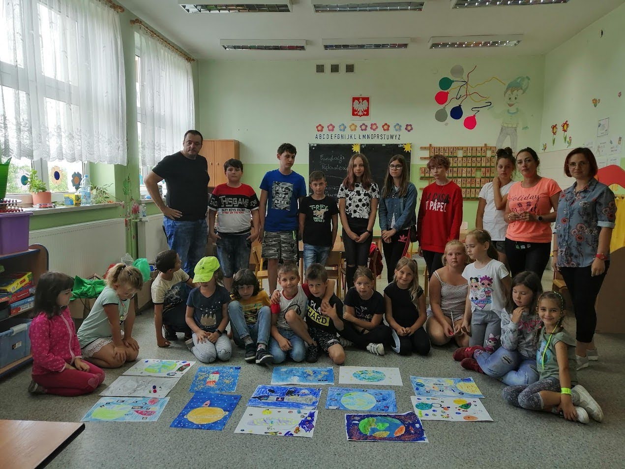 Grupa dzieci i dorosłych w sali lekcyjnej, prezentująca prace plastyczne ułożone na podłodze.