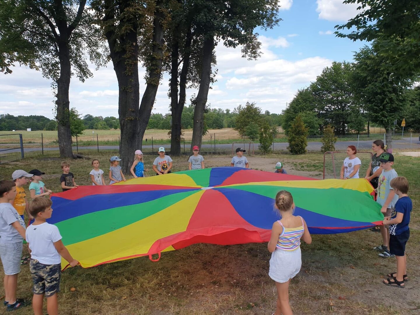 Dzieci stojące w kręgu na świeżym powietrzu trzymające kolorową chustę animacyjną.