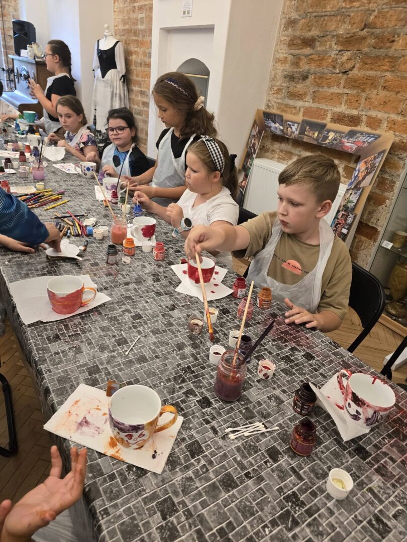 Grupa dzieci malująca ceramiczne kubki podczas warsztatów plastycznych przy długim stole.