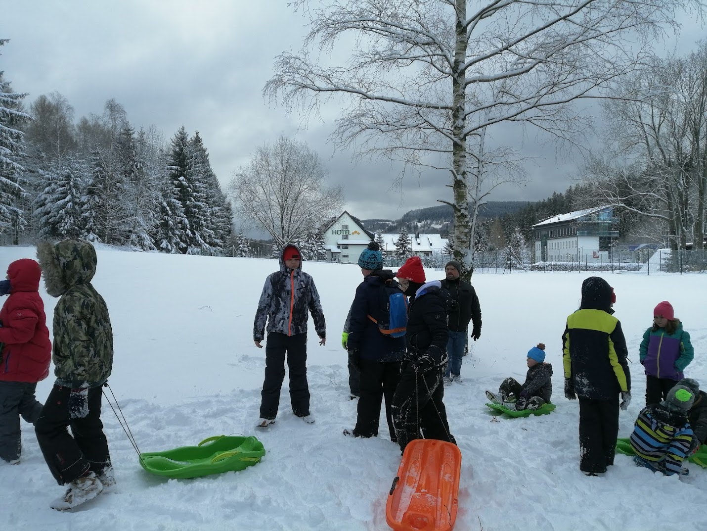 Dzieci i młodzież bawią się na stoku narciarskim, część z nich zjeżdża na sankach, inni przygotowują się do jazdy.