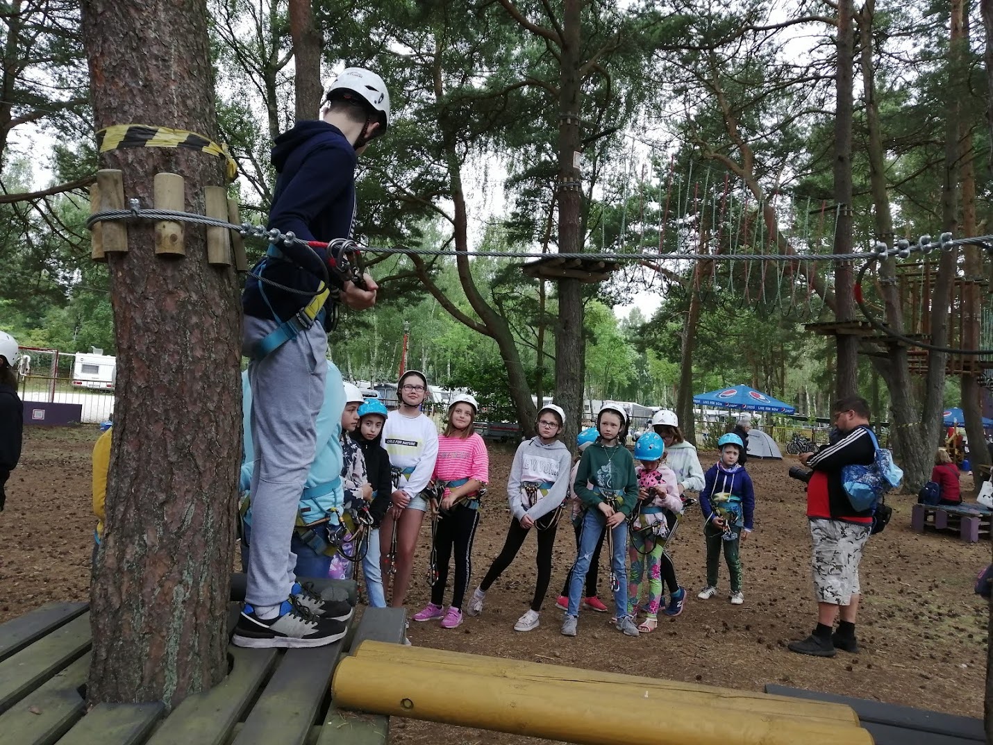 Grupa dzieci w uprzężach i kaskach czeka na instruktaż przy parku linowym podczas kolonii.