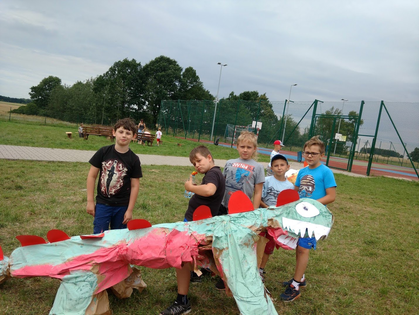 Grupa chłopców prezentuje papierowego smoka wykonanego podczas zajęć plastycznych na boisku.