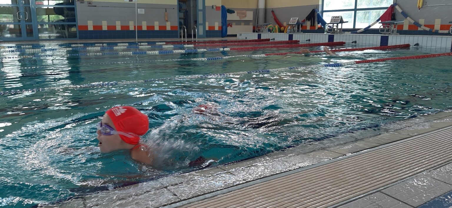 Dziewczynka w czerwonym czepku pływa stylem klasycznym w basenie podczas zajęć nauki pływania.